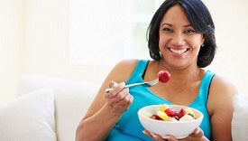 Overweight Woman Sitting On Sofa Eating Bowl Of Fresh Fruit Overweight Woman Sitting On Sofa Eating Bowl Of Fresh Fruit